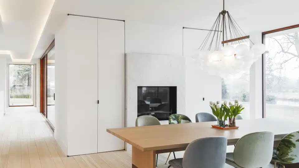 Moderne eetkamer met strakke afwerking: wit gestucte muren, naadloos schilderwerk, een houten eettafel en grijze gestoffeerde stoelen onder een opvallende bolvormige kroonluchter, met natuurlijk licht door grote ramen met zicht op groene omgeving. Pleisterwerk door In't Wit uit Herzele. Artikel in Paint & Stuc Magazine.