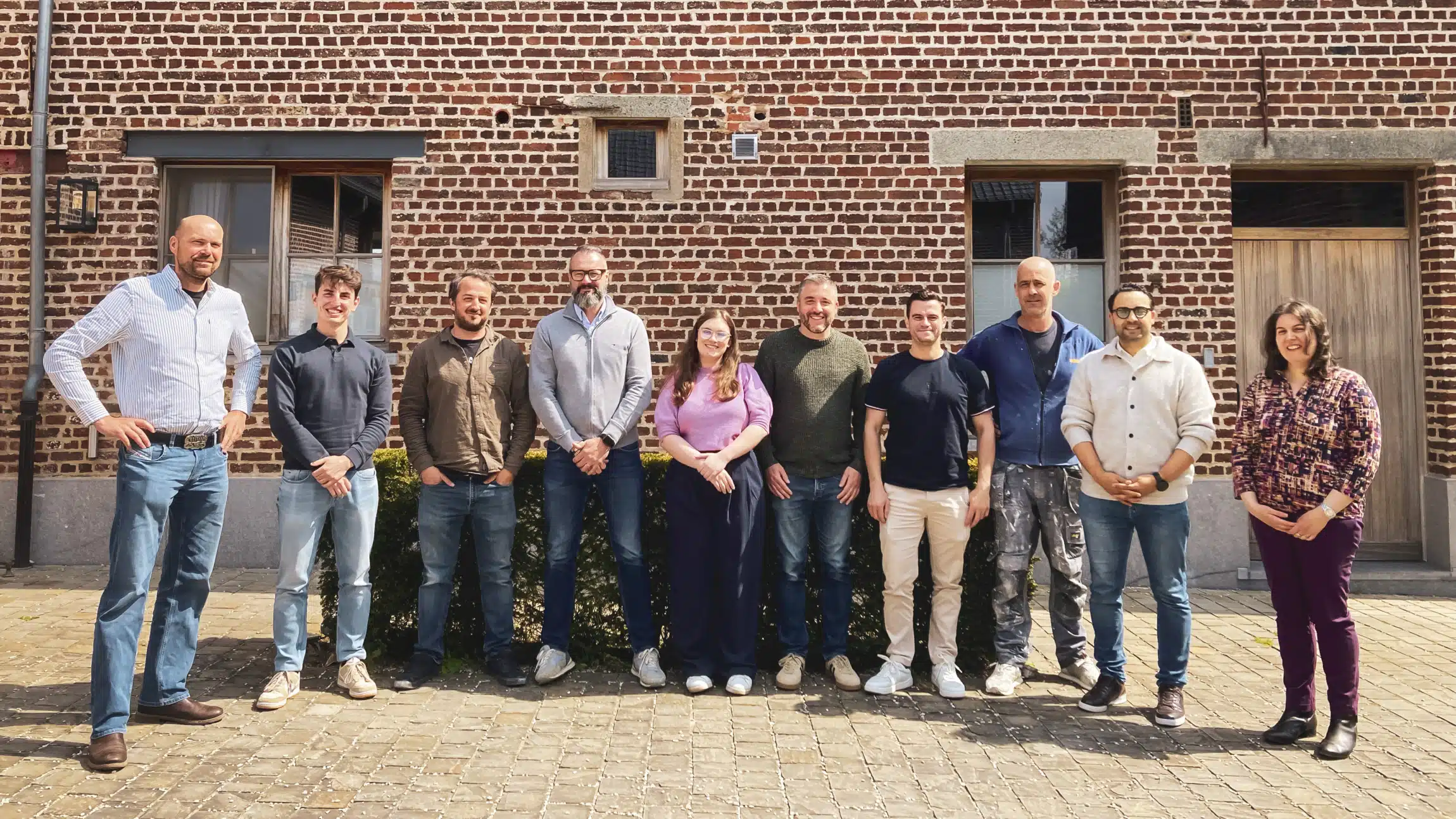 Team foto van In't Wit pleisterwerkbedrijf met tien medewerkers poseren voor bakstenen gebouw