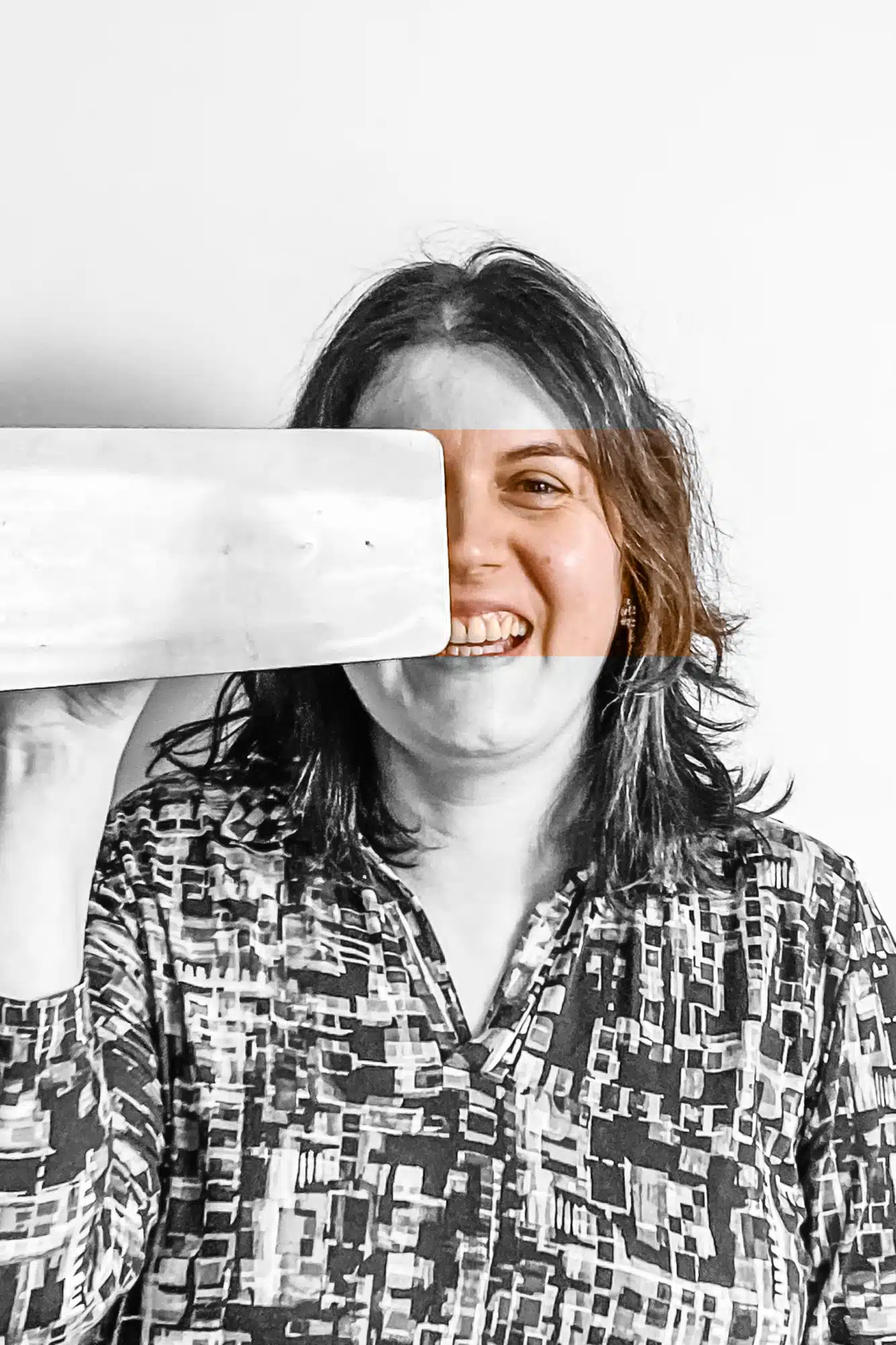 Vrolijke vrouw met donker haar houdt witte baksteen voor haar gezicht en lacht, draagt grijs geruit overhemd tegen witte acht
