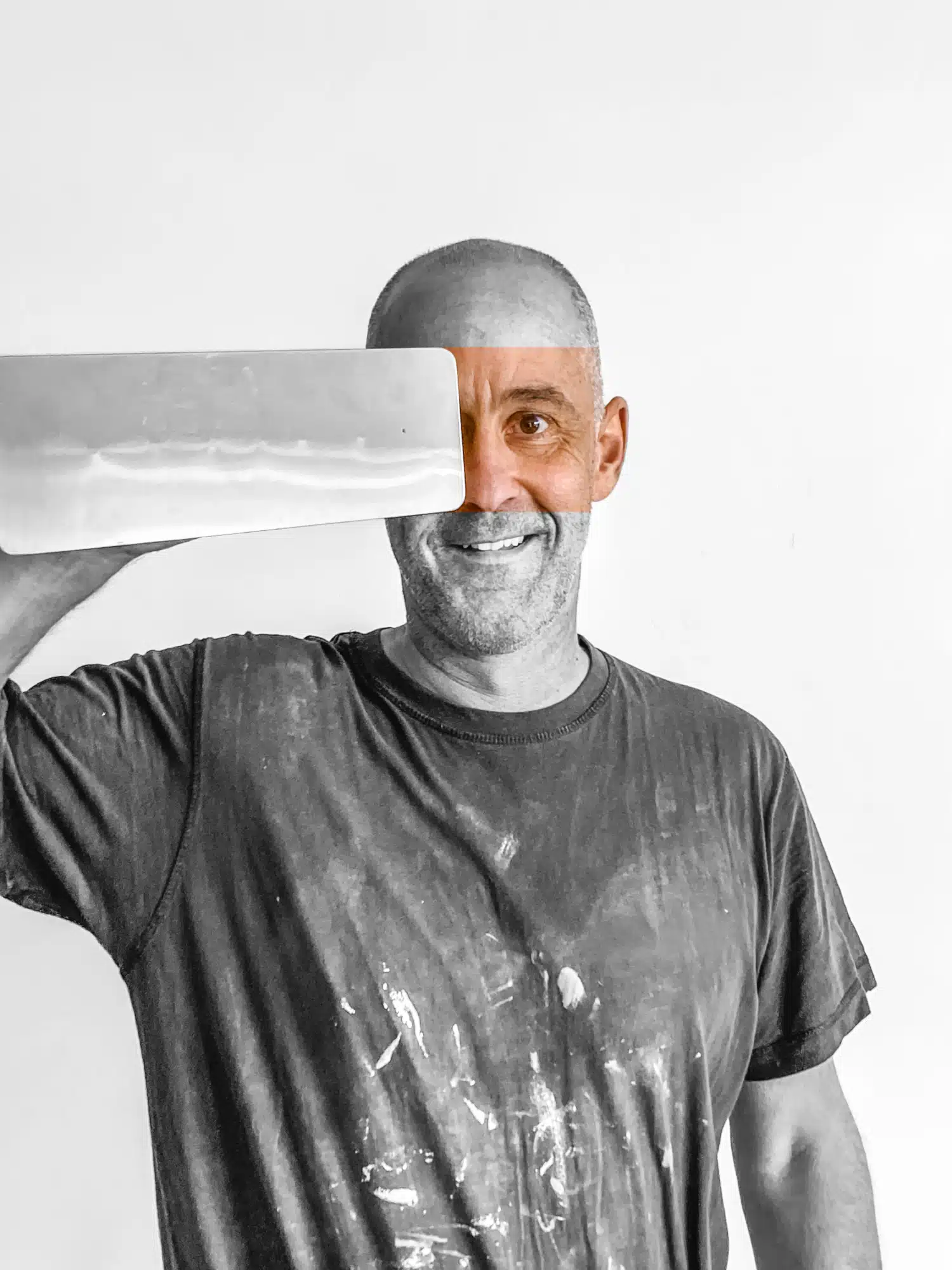 Glimlachende pleisterwerker met verfvlekken op shirt houdt plamuurmes omhoog, zwart-wit foto met kleuraccent op gezicht