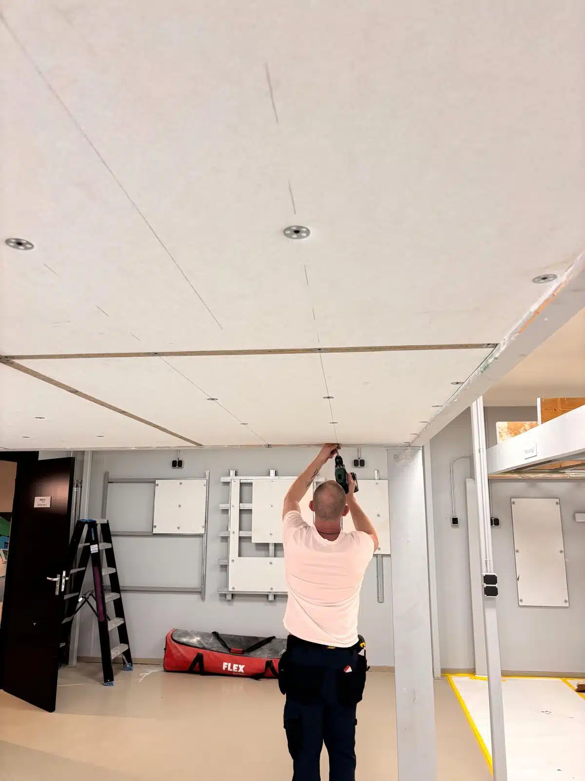 Vakman van In't Wit plaatst Eleganza plafondplaten met een boormachine tijdens renovatieproject, ladder en gereedschapstas op